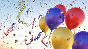A bundle of metallic colored balloons flying through many colorful confetti flakes and swirled ribbons. The whole scene is situated on a clear sky background.