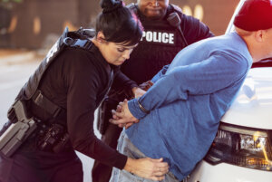 Cropped view of a policewoman and her African-American partner arresting a man. The suspect is leaning on the hood of a police car with his hands handcuffed behind his back. She is removing something from his pocket.