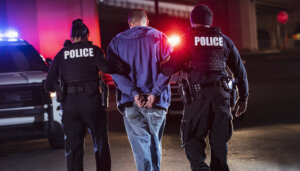Rear view of two multiracial police officers arresting a criminal suspect, a man with his hands handcuffed behind his back. One of the officers is an African-American man and the other is a policewoman, both in their 40s.