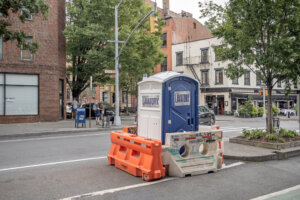 A portable plastic toilet for hardhat workers placed in the street surrounded with water filled plastic traffic barriers