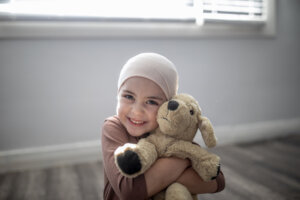 A sweet little girl battling cancer sits at home on the floor of her bedroom, with her arms wrapped tightly around her stuffed animal as she smiles gently for the portrait. She is dressed casually and wearing a headscarf as the sun shines in on her through the window.