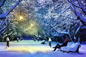 Beautiful scene in winter park at night with a teenage boy sitting on the bench and looking at the light of the street lamps.